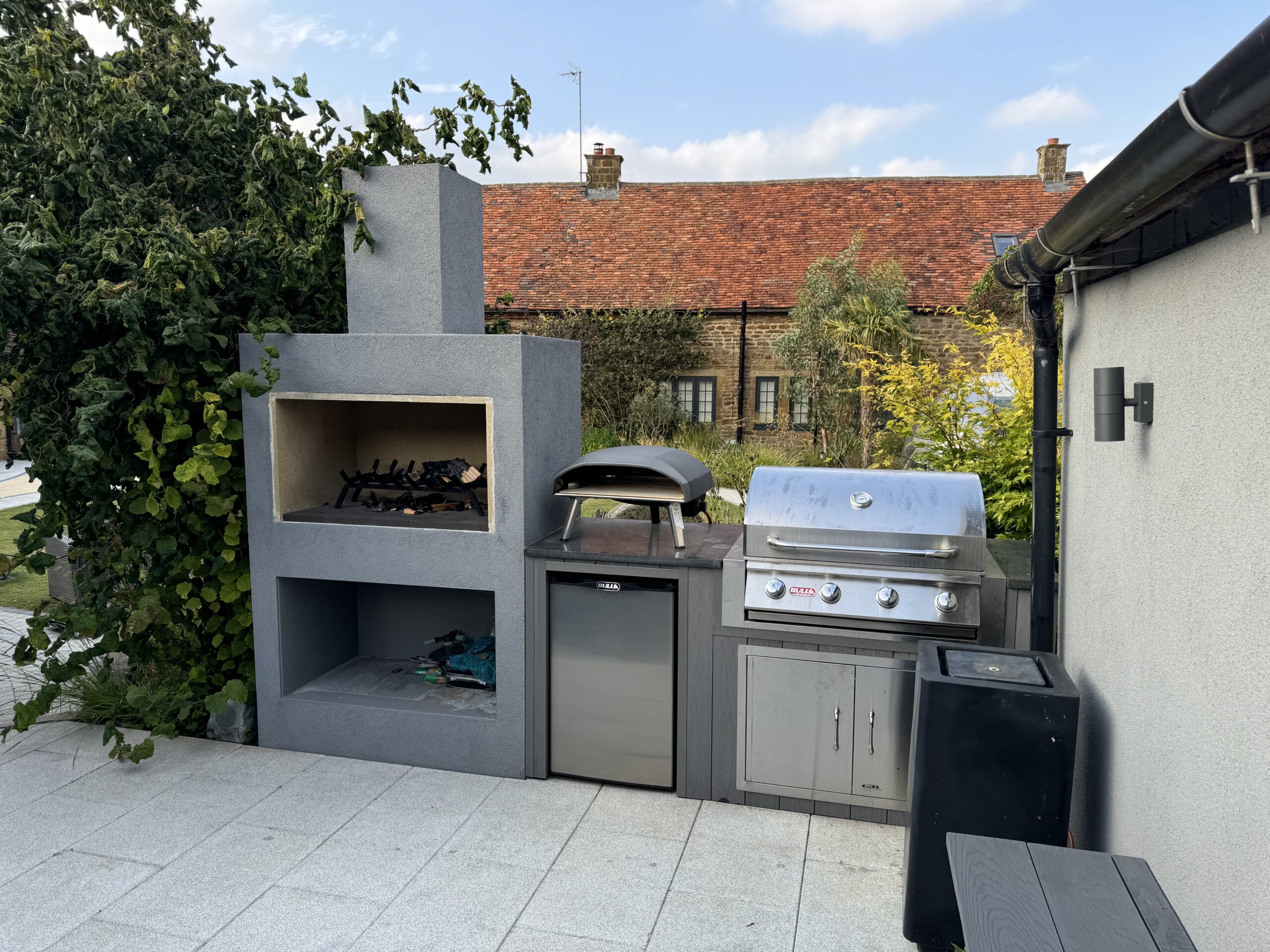 Grey rendered outdoor kitchen with pizza overn, log store, bbq, fire place and fridge.
